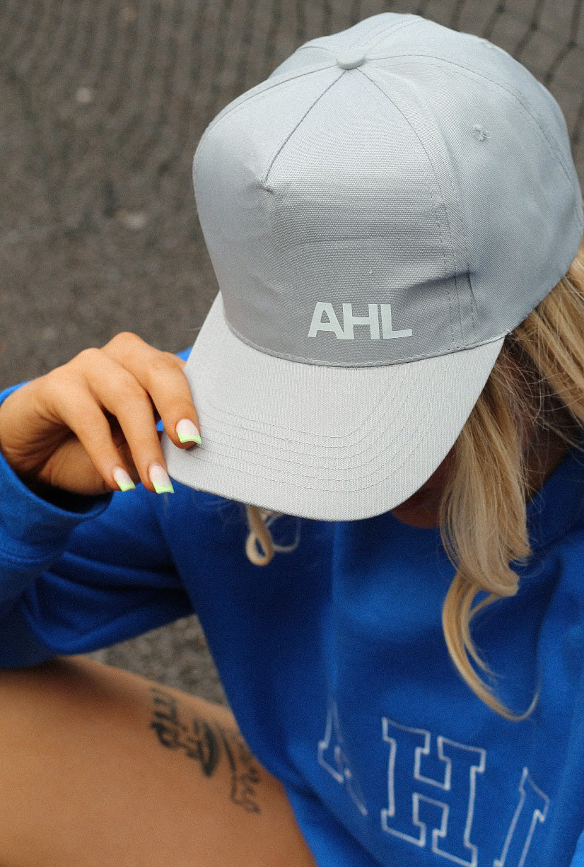 Female sitting on tennis court, looking down, whilst modelling grey AHL cap and blue sweatshirt