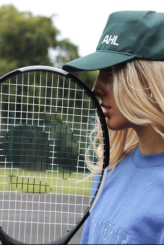 Female leaning tennis racket against face whilst wearing a khaki AHL cap