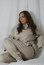 Load image into Gallery viewer, Female on sofa with white curtain in the background, modelling cream tracksuit
