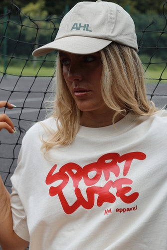 Female modelling cream Sport Luxe t-shirt whilst leaning against tennis net