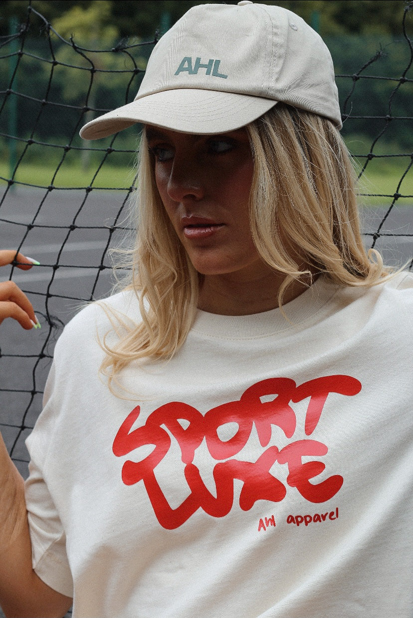 Female modelling cream Sport Luxe t-shirt whilst leaning against tennis net