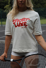 Load image into Gallery viewer, Female standing in front of tennis net, holding a tennis racket, modelling cream Sport Luxe t-shirt
