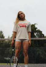 Load image into Gallery viewer, Female standing in front of tennis net with a tennis racket, wearing cream Sport Luxe t-shirt
