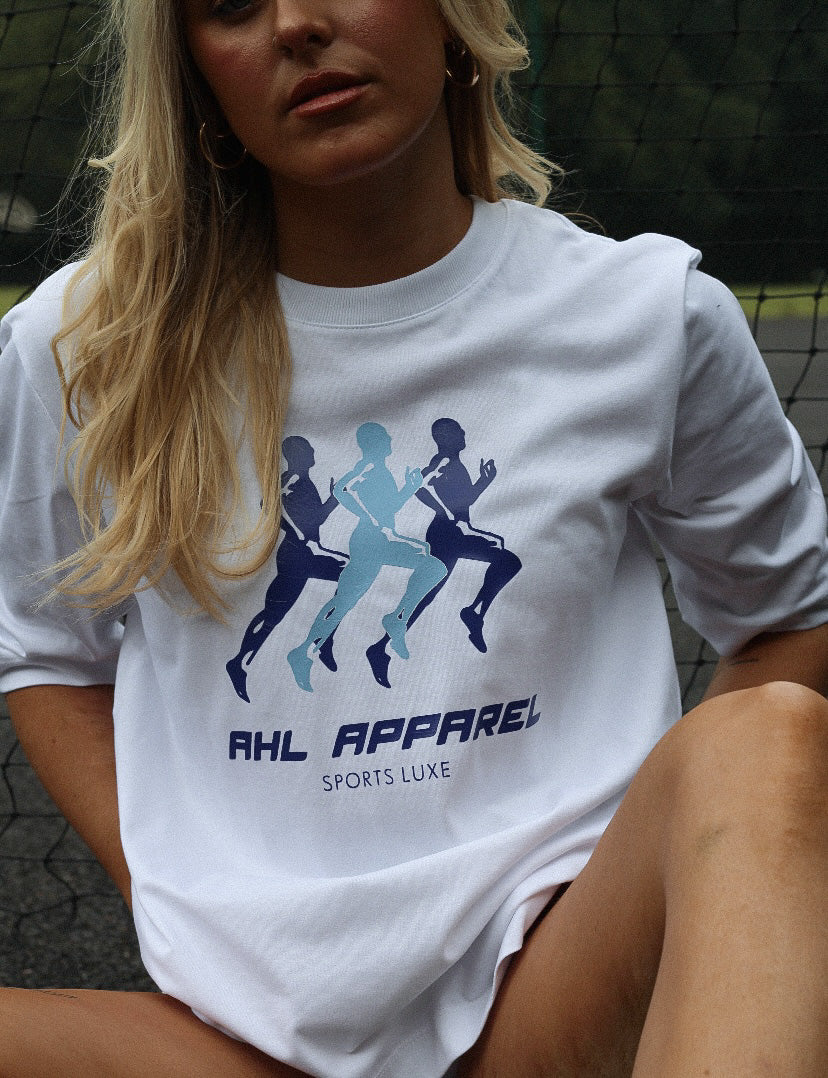 Female leaning against tennis net whilst modelling white Sport Luxe t-shirt