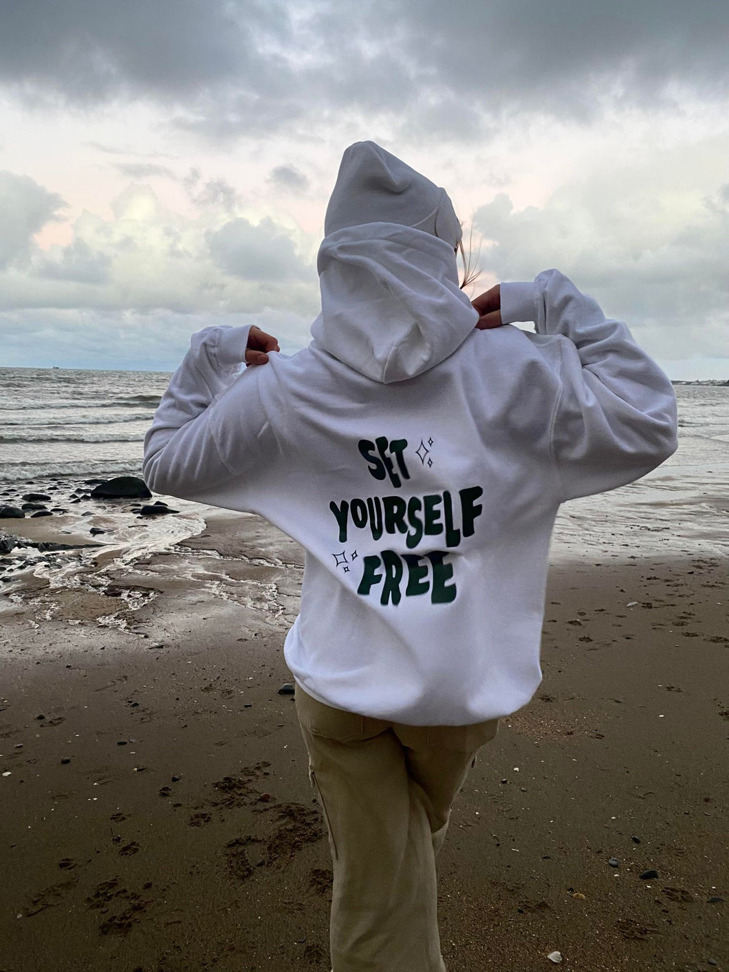 female modelling set yourself free hoodie and beanie in white on a beach