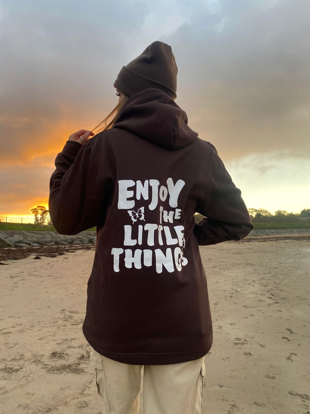 Female modelling brown enjoy the little things hoodie and beanie on a beach